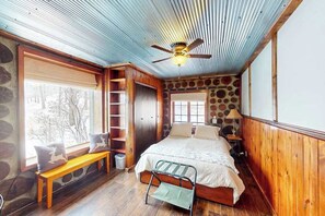 A bedroom with a bed, a bench, a ladder, a ceiling fan, and a window with a view of trees.