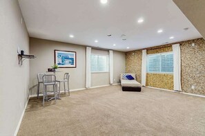 Ein Zimmer mit Teppichboden, einem Bett mit Kissen, einem Schreibtisch mit Stühlen, einem gerahmten Bild an der Wand und einem Fenster mit Jalousien.