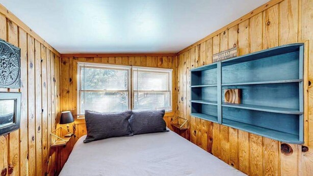Ein Zimmer mit Holzwänden, einem Bett mit weißer Bettwäsche, zwei Nachttischlampen und einem Fenster mit Jalousien.