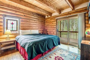 Quarto de cabana de tronco com uma cama, uma mesinha de cabeceira, uma luminária, uma janela com design em vitral e uma porta de correr que dá para um deck externo.