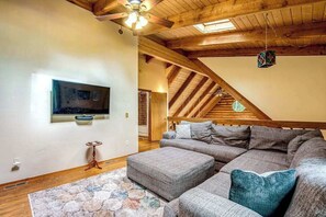 Uma sala de estar com um amplo sofá modular, uma televisão de tela plana fixada na parede e teto de madeira com vigas aparentes.