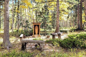 Een boslandschap met een houten constructie die lijkt op een schrijn of altaar, omringd door bomen en een stenen bank.