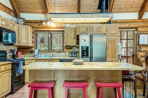 A kitchen with wooden cabinets, a central island with stools, a refrigerator, and a microwave. - Pikes Peak Retreat - Cabin w/ Pikes Peak & Forest Access (Divide)
