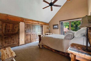 A bedroom with a wooden bed frame, a ceiling fan, and a large window with a view of greenery outside. - Reunion Lodge • Hot Tub, Game Room & Views (Sturgis)