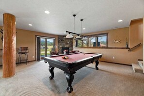 A pool table in a room with a stone wall, a fireplace, and a view of the outdoors. - Reunion Lodge • Hot Tub, Game Room & Views (Sturgis)