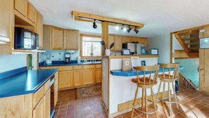 A kitchen with wooden cabinets, a blue countertop, a coffee machine, and a bar area with stools. - Friends & Family Base Camp - Dog-Friendly 3-Story Group Getaway (Woodland Park)