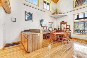 A spacious room with a wooden floor, a dining table with chairs, a kitchen area with a counter, and large windows providing natural light. - Boulder Ridge Lodge - Hot Tub Log Cabin Near Golf Course (Lead)