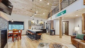 A spacious living area with a high ceiling, a kitchen with modern appliances, a dining area with a large window, and a cozy seating area with a fireplace. - Sundby Cabin - New Chalet • Walk to Skiing (Lead)
