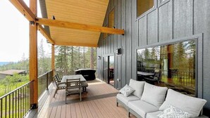 A wooden deck with a sofa and table, overlooking a forested area. - Sundby Cabin - New Chalet • Walk to Skiing (Lead)