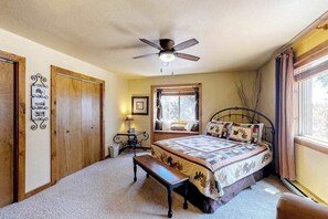 Une chambre à coucher comprenant un lit, un ventilateur de plafond, une fenêtre avec des rideaux, un banc et un cadre photo fixé au mur.