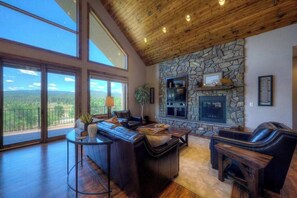 A spacious living room with a stone fireplace, leather furniture, and large windows offering a view of the outdoors.