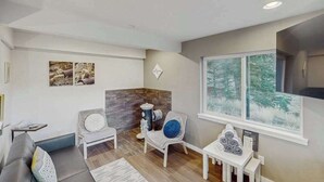 A modern living room with a grey sofa, a white armchair, a small table with a blue pillow, and a window with a view of trees. - Mtn-View Apt • Private Hot Tub, Dogs OK (Green Mountain Falls)