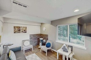 Ein modernes Wohnzimmer mit einem grauen Sofa, einem weißen Sessel, einem kleinen Tisch mit einem blauen Kissen und einem Fenster mit Blick auf Bäume.