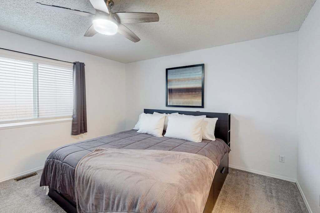 A bedroom with a bed, a ceiling fan, a window with curtains, and a wall-mounted painting.