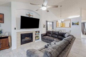 Ein Wohnzimmer mit Kamin, einem großen Flachbildfernseher, einem grauen Sofa und einem Deckenventilator.