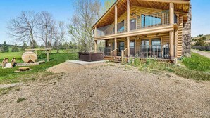 A two-story log cabin with a balcony, a hot tub, and a gravel driveway. - Waterwheel Retreat - Classic Home • Hot Tub & Cozy Loft (Sturgis)