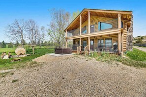A two-story log cabin with a balcony, a hot tub, and a gravel driveway. - Waterwheel Retreat - Classic Home • Hot Tub & Cozy Loft (Sturgis)