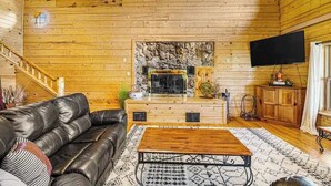 A wooden cabin interior with a stone fireplace, leather sofa, and a flat-screen TV. - Waterwheel Retreat - Classic Home • Hot Tub & Cozy Loft (Sturgis)