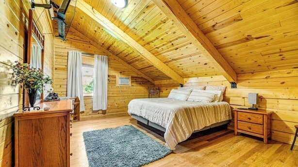 A cozy wooden cabin bedroom with a bed, bedside tables, a dresser, and a window with curtains. - Waterwheel Retreat - Classic Home • Hot Tub & Cozy Loft (Sturgis)