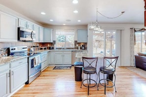 A kitchen with white cabinets, a microwave, and a stove. There are two bar stools and a trash can. - Windy Hill Retreat - Dog-Friendly Home • Huge Fenced Yard (Colorado Springs)