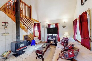 A living room with a fireplace, a sofa, a television, and a staircase. - Pine Perch - Cozy A-Frame • Wood Stove & Balcony (Florissant)
