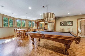 A pool table in a room with wooden flooring, a dining area with chairs, and a kitchen with wooden cabinets. - Eagle Crest Nest - Secluded Cabin • Hot Tub & Decks (Spearfish)