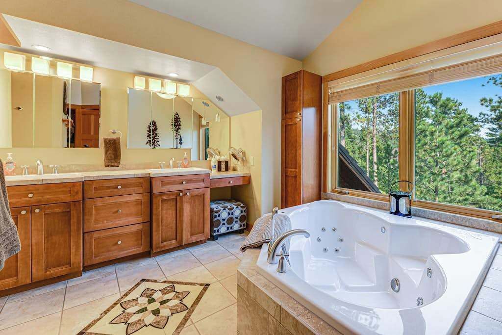 Ein modernes Badezimmer mit einer großen Badewanne, Holzschränken, einem beleuchteten Spiegel und Blick auf Bäume durch ein Fenster.