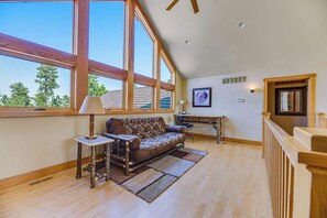 A spacious room with a high ceiling, large windows, a sofa, a table, and a lamp. - Eagle Crest Nest - Secluded Cabin • Hot Tub & Decks (Spearfish)