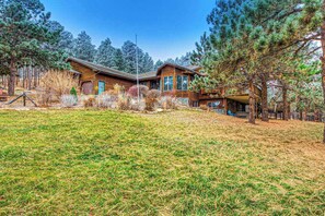 A two-story log cabin with a large front porch, surrounded by a grassy area and pine trees. - 20-Acre Lodge • Hot Tub, Sauna • Dogs OK (Rapid City)