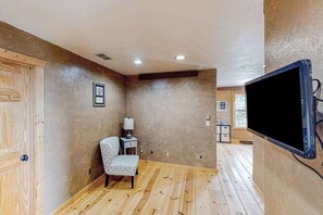 Une pièce avec un plancher en bois, un téléviseur fixé au mur, une chaise et une petite table avec une lampe.