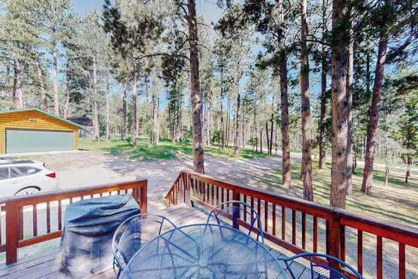 A wooden deck with a metal table and chairs, overlooking a forested area with a garage and a car parked nearby. - Mountain Escape - Mountain-View Home + Cabin w/ Deck (Sturgis)