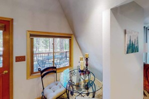 A room with a glass table, a cushioned chair, a window with a view of outdoor furniture, and a painting on the wall. - Mountain Escape - Mountain-View Home + Cabin w/ Deck (Sturgis)