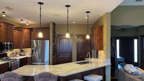 Una cocina con isla central, electrodomésticos de acero inoxidable, gabinetes de madera e iluminación colgante, y vistas al área de estar.