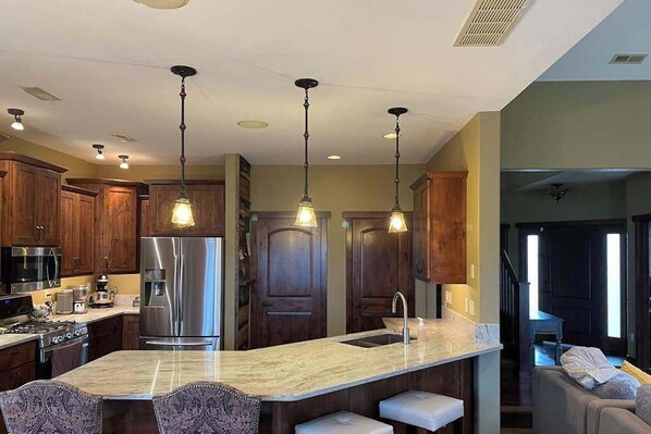 A kitchen with a central island, stainless steel appliances, wooden cabinets, pendant lighting, and a view into the living area. - Big 4BR Near Mt Rushmore • Fireplace • Rapid City (Rapid City)