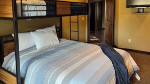 Una cama litera con una cama plana en la parte inferior, un televisor montado en la pared y una puerta que da a otra habitación.
