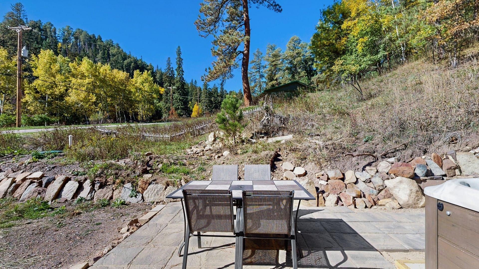 Une terrasse extérieure avec une table et des chaises, entourée d’un mur de pierre et bordée par une colline boisée.
