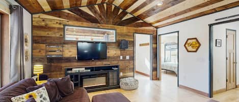 A modern living room with a wooden ceiling, a flat-screen TV mounted above a fireplace, and a sofa with cushions.