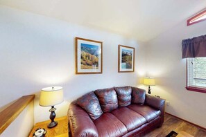 A living room with a brown leather sofa, two framed pictures on the wall, a lamp on a side table, and a window with blinds. - Horseshoe Haven - Mountain-View Home • Deck & Grill (Divide)