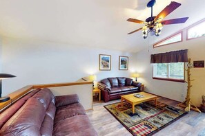 A living room with a brown leather sofa, a wooden coffee table, a ceiling fan, and a window with curtains. - Horseshoe Haven - Mountain-View Home • Deck & Grill (Divide)