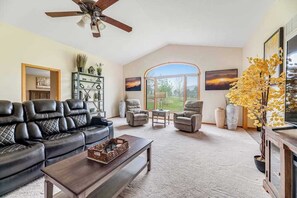 A spacious living room with a black leather sofa, a coffee table, and a fireplace. There are two armchairs and a side table. The room has a large window with a view of the outdoors.