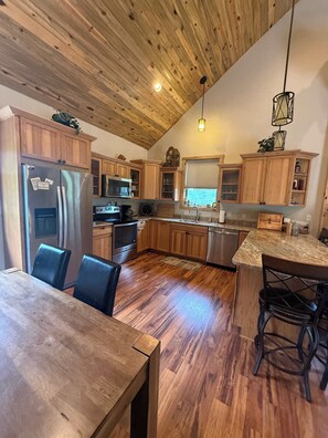 A kitchen with wooden cabinets, a stainless steel refrigerator, a built-in oven, and a dining area with a wooden table and chairs. - Legacy Lodge- 4BR Lodge Minutes from Deadwood (Sturgis)