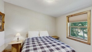 A bedroom with a bed, a nightstand, a lamp, and a window with a view of trees. - Legacy Lodge - 4BR Lodge Minutes from Deadwood (Sturgis)