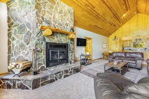 A cozy living room with a stone fireplace, leather furniture, and a kitchen in the background. - Lazy Bear Lodge - Rustic Lodge • Hot Tub & Wrap Deck (Sturgis)