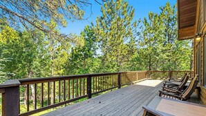 A wooden deck with a railing, surrounded by trees, and a building with a sloped roof. - Lazy Bear Lodge - Rustic Lodge • Hot Tub & Wrap Deck (Sturgis)