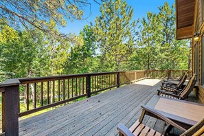 A wooden deck with a railing, surrounded by trees, and a building with a sloped roof. - Lazy Bear Lodge - Rustic Lodge • Hot Tub & Wrap Deck (Sturgis)