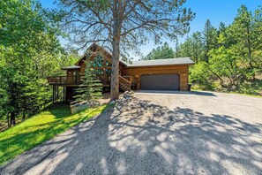 A cabin with a deck, a garage, and a gravel driveway surrounded by trees. - Lazy Bear Lodge - Rustic Lodge • Hot Tub & Wrap Deck (Sturgis)