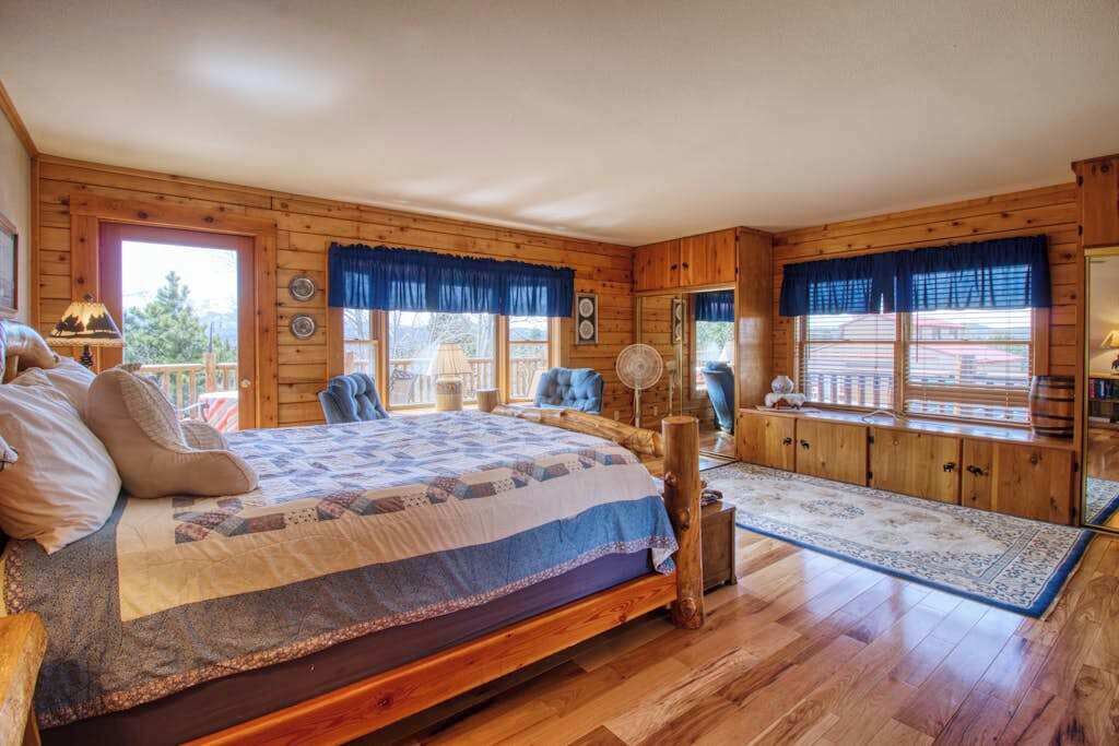 A wooden cabin bedroom with a large bed, a fan, and a view of the outside.