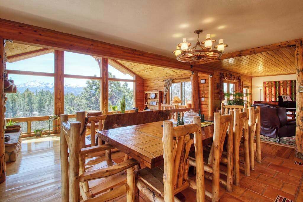 Interior de madera con una gran mesa de comedor y sillas, una araña y vistas al exterior.