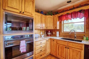 A kitchen with wooden cabinets, a stainless steel oven, and a window with a red valance. - Mills Ranch - Hot Tub Cabin • Pikes Peak Views (Woodland Park)