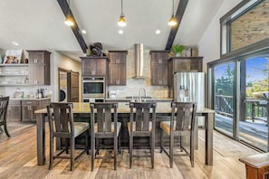 A modern kitchen with dark wood cabinets, a central island with bar stools, and a large window providing a view of the outdoors. - Pillar Peak Perch - Luxury Lodge • Hot Tub & Big Views (Sturgis)